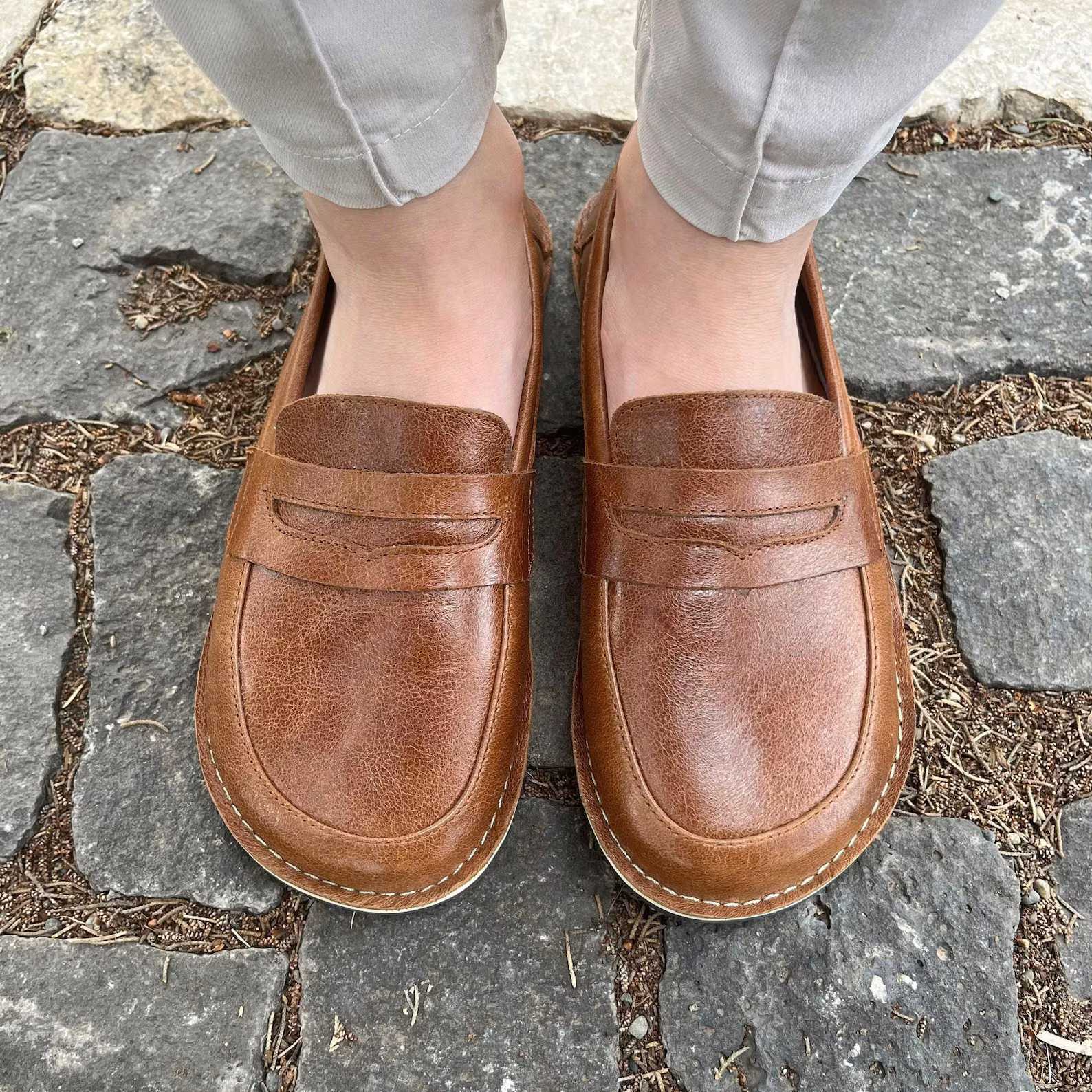 A top down view of a pair of feet standing on cobbletstones wearing brown leather wide toe box barefoot penny loafers