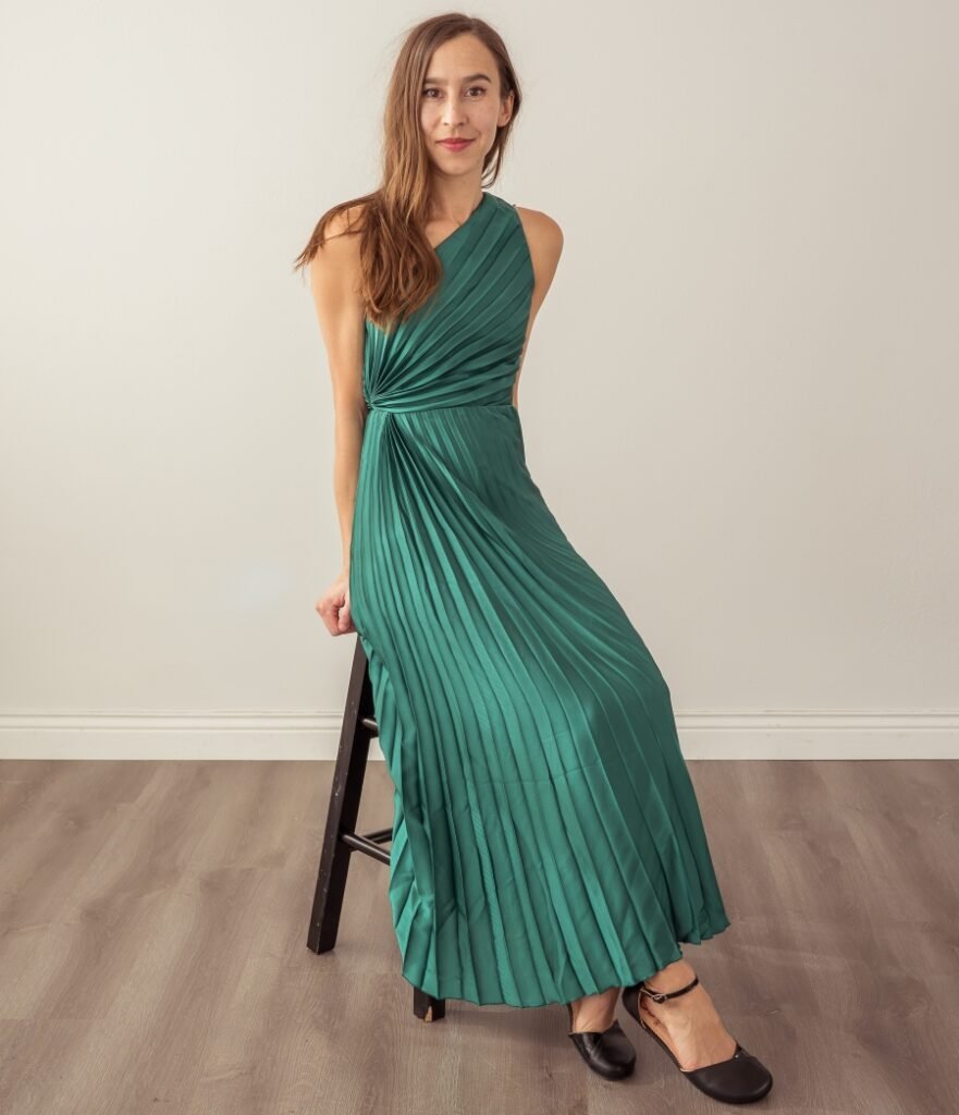 A woman with long brown hair wearing a one shouldered green pleated Abercrombie and Fitch dress leaning against a stool with a slight smile on her face. On her feet are wide toe box dressy barefoot flats Shapen Poppy with black leather.
