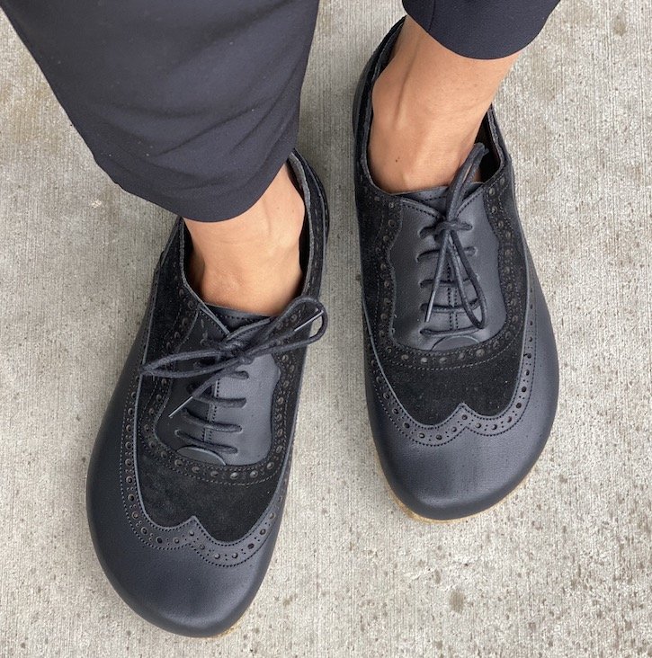 a top down view of a pair of feet standing on concrete wearing black oxfords with brogue detailing in a natural foot shape