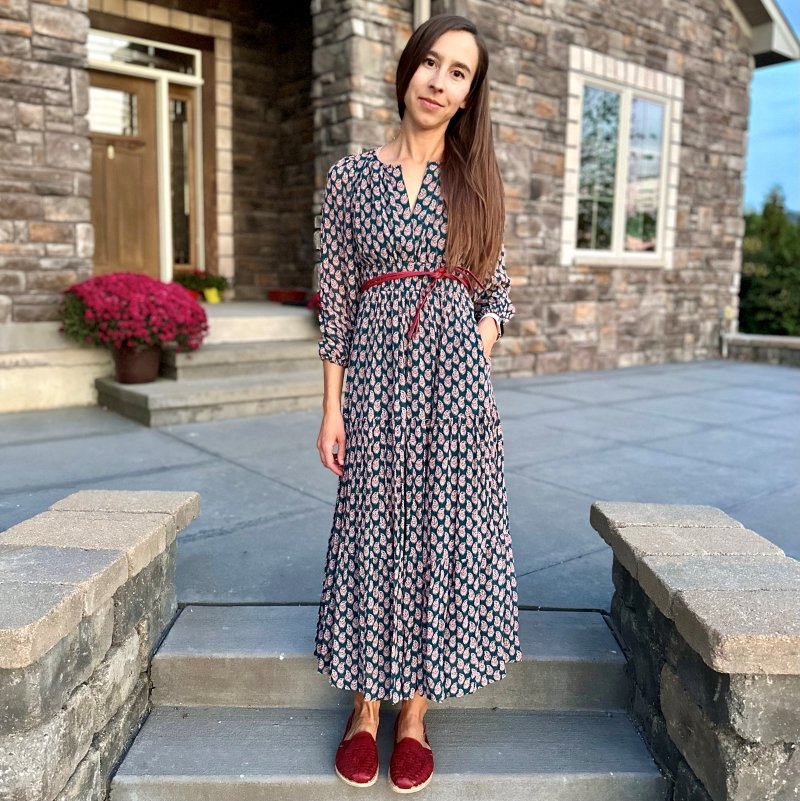 A woman with long brown hair pulled to one side standing on her front porch steps wearing an long printed Anthropologie dress and Origo Huarache Slip ons by Anya in a red color with matching leather belt