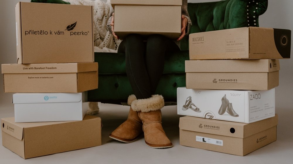 A stack of shoe boxes from various brands stacked up in front of a green velvet couch where a person sits wearing Zeazoo Dingo boots in light brown