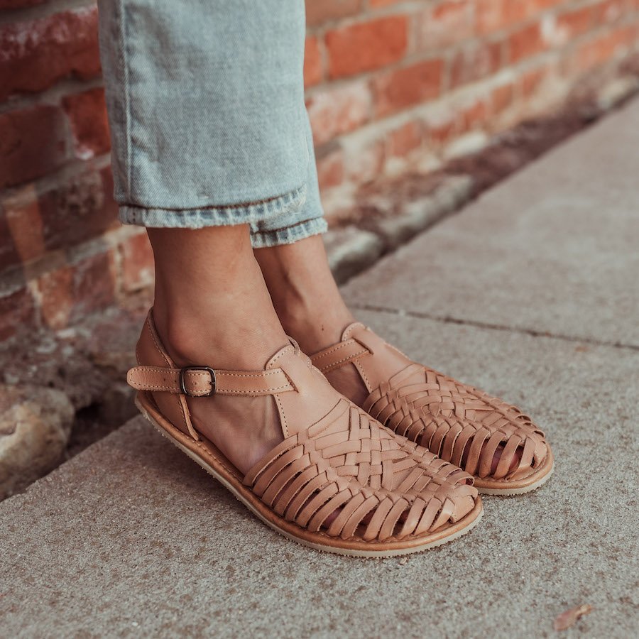 a close up of a pair of feet wearing tan wide toe box huaraches with a natural foot shape and a buckle around the ankle