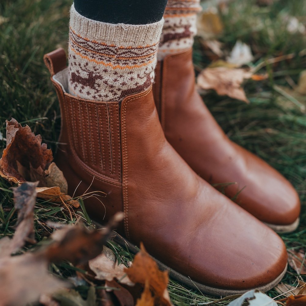 A side angled view of a pair of feet standing in grass wearing brown Lila barefoot chelsea boots, designed by Anya and sold at Anya's Shop with a pair of cozy wool socks peeking out the top