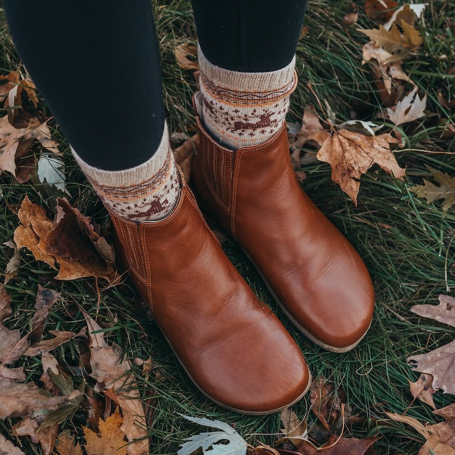 Front angled view of Anya's Shop exclusive Lila Chelsea boots in a warm brown leather with leather covered elastic. The person is standing on leaf strewn grass with fall themed sock poking out the top of the boots. The wide box is featured in the forefront of the image
