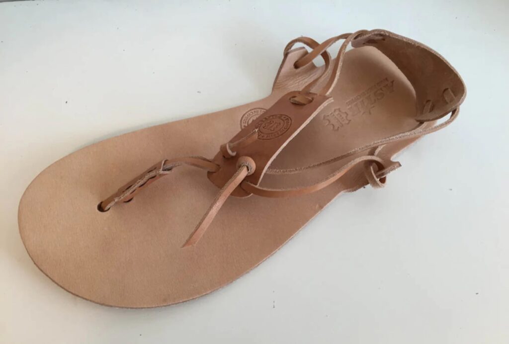A tan leather grounding sandal with thin leather straps that wrap around the sandal shown on a white background. Sandals have a rectangular leather heel grab on the back of the shoe straps and a similar one on the top of the foot.