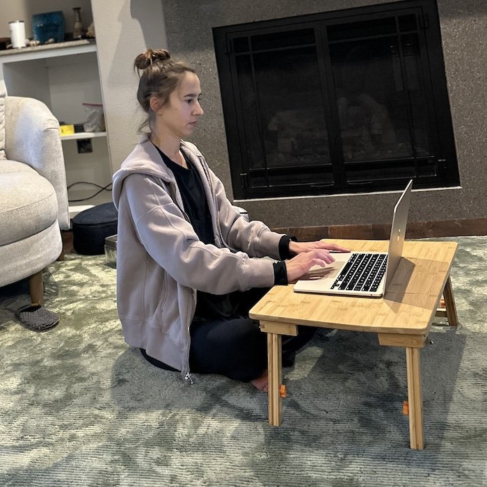 A woman sitting on the floor cross legged at a bamboo laptop tray with a laptop on top