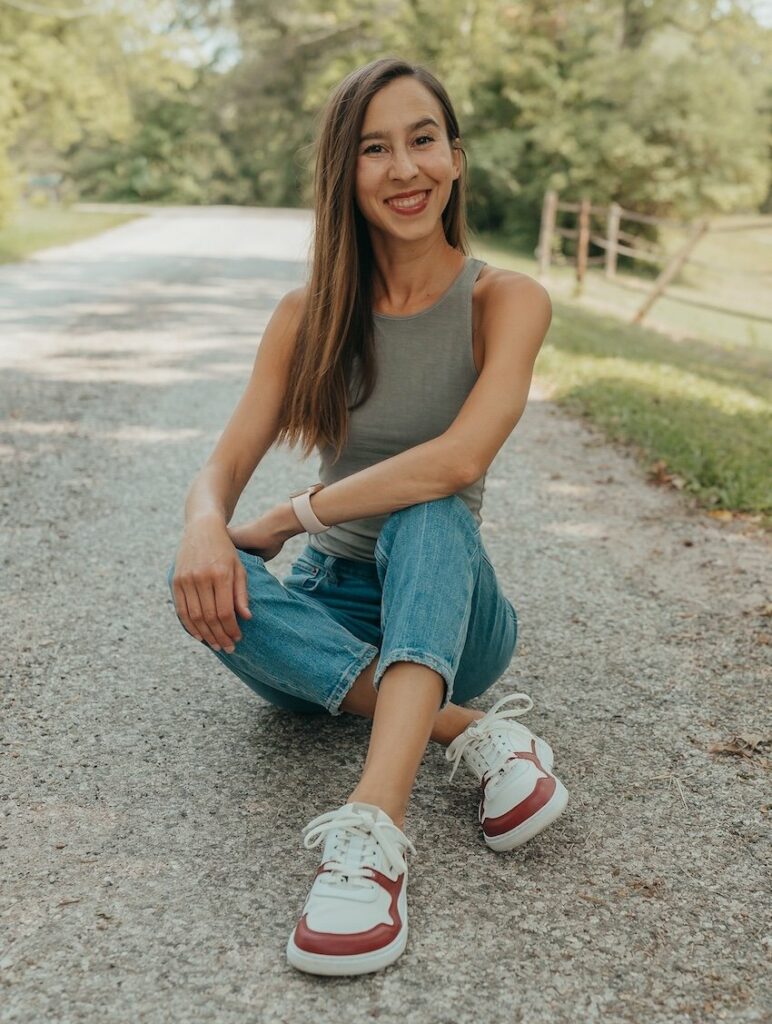 A woman sitting on the ground with legs crossed smiling at the camera, wearing a tank, jeans, and custom burgundy Barebarics X Anya Zing barefoot sneakers with a wide toe box and anatomical shape