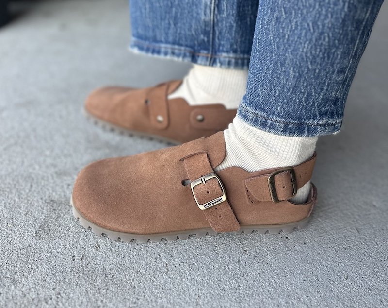 Close up side view of a person's feet in Barebound Ikarus barefoot clogs with a wide toe box and flexible outsole. The side profile shows the flat outsole well, and the model has worn them with light colored socks and denim.