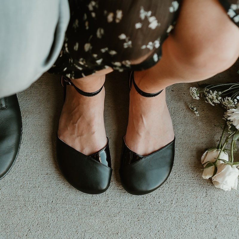 A top down view of a woman's feet with the hem of her dress and flower bouquet visible around the edges - she is wearing black leather Shapen Poppy barefoot dress sandals that can replace high heels