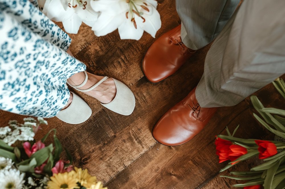 Top down view of a man and a woman dressed up in fancy attire focusing on their feet. The woman is wearing light colored Shapen Orchid barefoot flats