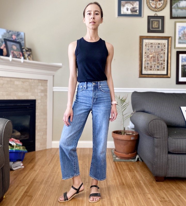 A woman standing inside wearing a tank top and jeans and black Be Lenka Barefoot Summer Sandals made of leather