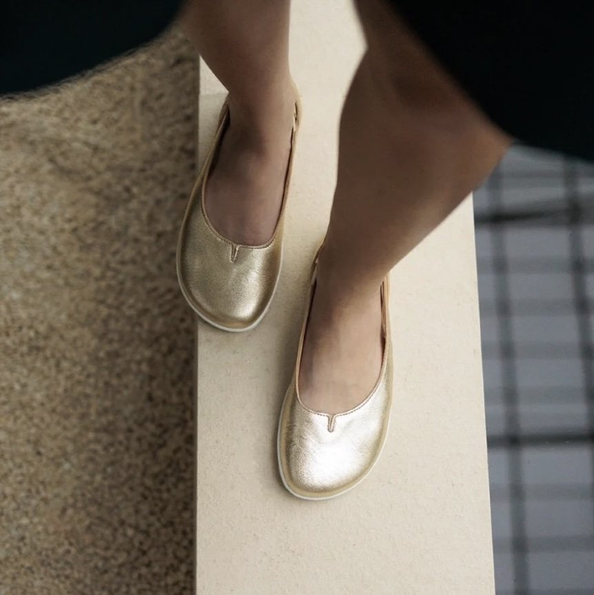 A top down view of Be Lenka Bellisima barefoot fancy flats in gold. One foot is in front of the other on a wide ledge, and the anatomical toe box is prominent