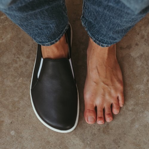 Top down view of a person with wide feet standing with one shoe on one shoe off. On the left is Be Lenka Eazy Neo leather barefoot slip-ons for wide feet