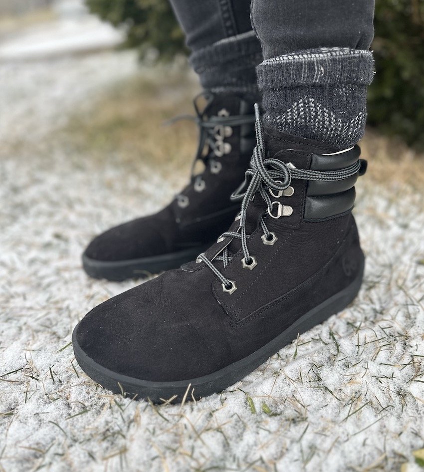 A close up of a pair of feet standing on snowy ground wearing Be Lenka Nevada barefoot boot in black leather