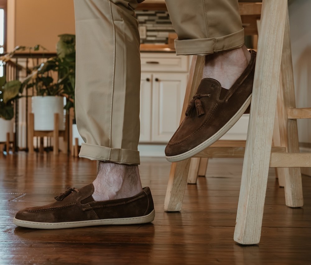 A close up of a man's feet and lower legs resting on a bar stool wearing tan pants and brown leather loafers with a tassel. They are Be Lenka Portsider barefoot loafers, with a flat sole and wide toe box.