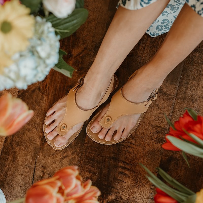 A top down view of a woman's feet resting on a wood floor with flowers visible all around and the hem of her dress. She is wearing Be Lenka Promenade sand leather barefoot sandals for women