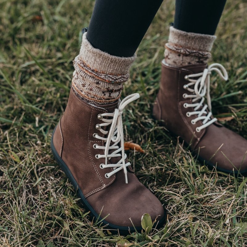 A stylized photo of Be Lenka Winter chocolate brown wide toe box boots for snow and cold. The person is wearing patterned chunky socks and black leggings
