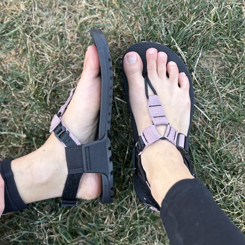 A pair of feet resting on grass outside wearing Bedrock Cairn Evo barefoot sandals with cushion with a lilac purple strap and vegan materials