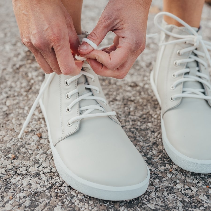 A close up of a pair of feet wearing Be Lenka Atlas barefoot boots in cream with a hand tying the laces