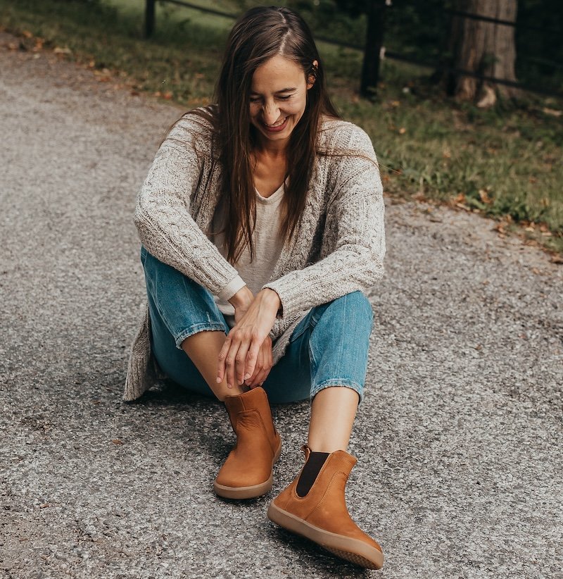 A woman with long brown hair wearing jeans and an oatmeal colored sweater sitting on the ground outside. She is looking down, cracking a smile, with her elbows on her knees. Be Lenka Entice barefoot chelsea boots in a light brown cinnamon color with a wide box are on her feet in the forefront of the image.
