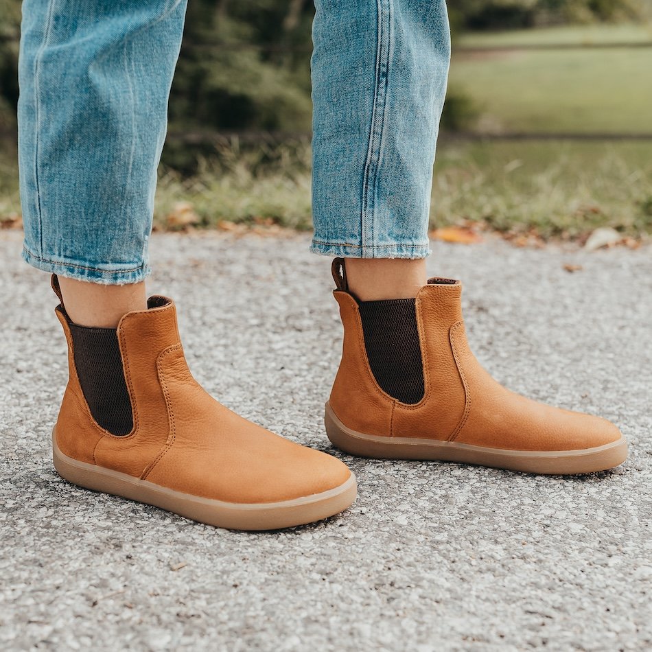 Side view of a person in light colored jeans wearing Be Lenka Entice barefoot chelsea boots in Cinnamon outside on gray pavement. Grass and trees are in the background.