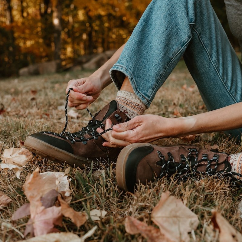 A person from the legs down sitting on grass with fall leaves around, wearing jeans, wool socks, and Be Lenka Ranger brown leather wide toe box barefoot hiking boots. Person is tying their shoelaces.