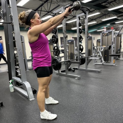 A full body photo of a woman in a pink top and black shorts wearing white canvas barefoot lifting shoes for the gym. She is swinging a kettle bell up in front of her face.