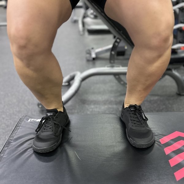Front close up of a pair of muscular woman's legs standing on a box in the gym wearing Notorious Lift barefoot zero drop wide toe box shoes in black for gym exercise
