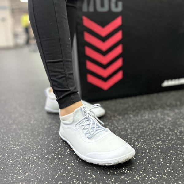 A front angled close up of a pair of woman's leg in black leggings at the gym wearing Tolos Archetype 2.0 barefoot gym shoes with a wide toe box in white.