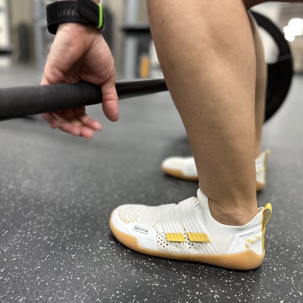 Close up side back view of TYR DropZero barefoot trainers being worn by a person lifting weights at the gym. The flat outsole with no heel is helping with balance.