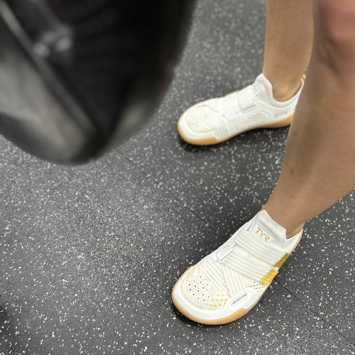 A top angled view of TYR DropZero barefoot wide toe box gym shoes in white with gold accents. A blurred weight is visible in the tope left of the image.