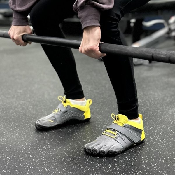 A woman in black leggings and Vibram Five Fingers athletic gym shoes being worn by a woman lifting weights at the gym. Her hands are gripping the bar and her feet are planted firmly on the ground.