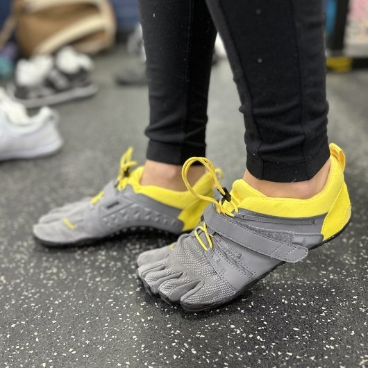 A side flexed view of a person's feet in Vibram Five Fingers on the gym floor.They are gray, have individual toe pockets for ultimate foot function, and yellow accents.