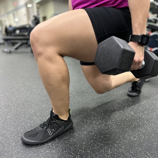 A waist down view of a person doing a weighted lunge wearing black Vivobarefoot gym shoes.