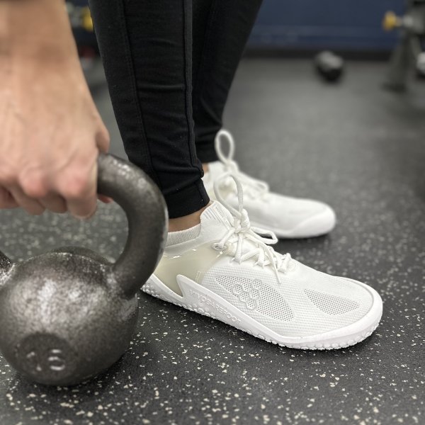 A side close up view of white vivobarefoot gym shoes with flat zero drop outsole being worn in the gym with the person about to pick up a 15lb kettlebell.