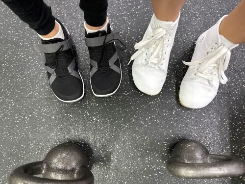 Top down view of 2 people standing side by side at the gym with kettlebells on the floor in front of their feet. The person on the left is wearing Xero Shoes, and the person on the right is in Bearfoot Ursus high top canvas in white. Both have a wide toe box.