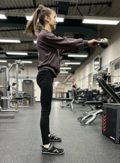 A full body view of a woman with a long brown ponytail, purple sweatshirts, and black leggings doing a kettle bell swing wearing Xero Shoes black barefoot gym shoes.