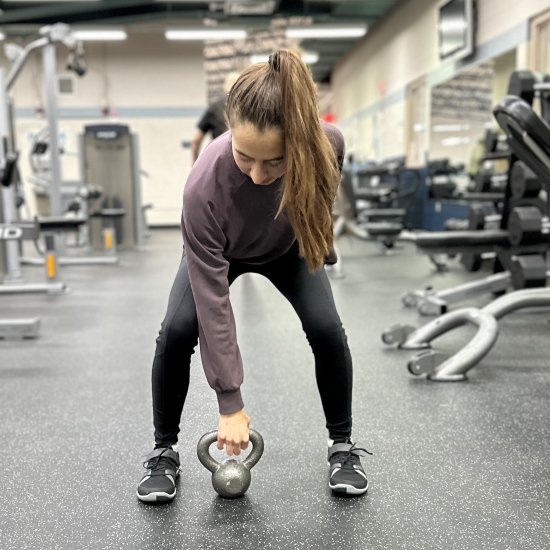 A woman with a long brown pony tail, purple sweatshirts, and black leggings squatting to pick up a kettlebell with her right hand. On her feet are Xero Shoes black and gray barefoot gym shoes for exercise