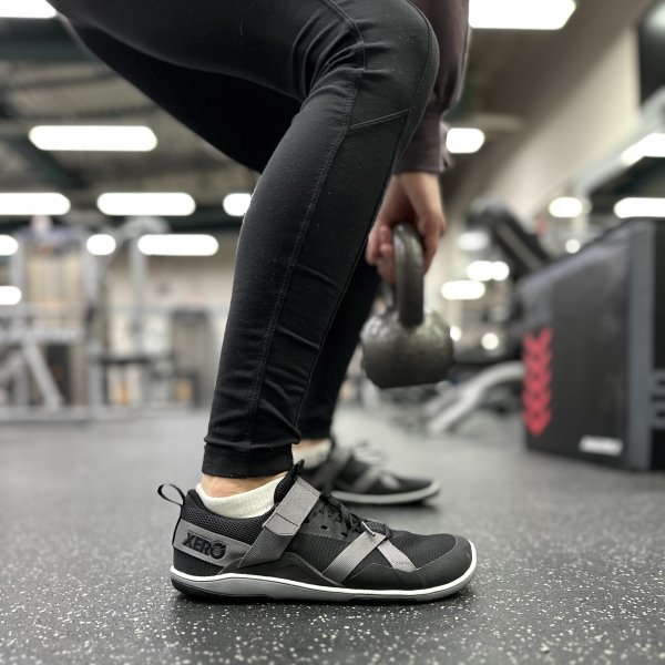 Side close up view of a person squatting with a kettle bell in their hands wearing Xero Shoes black barefoot zero drop athletic shoes for the gym