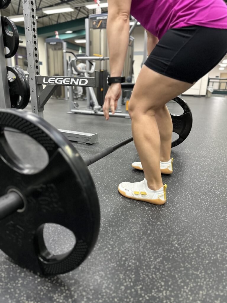 A woman bending over to pick up a barbell for a deadlift, weightlifting in the gym wearing TYR DropZero barefoot trainers