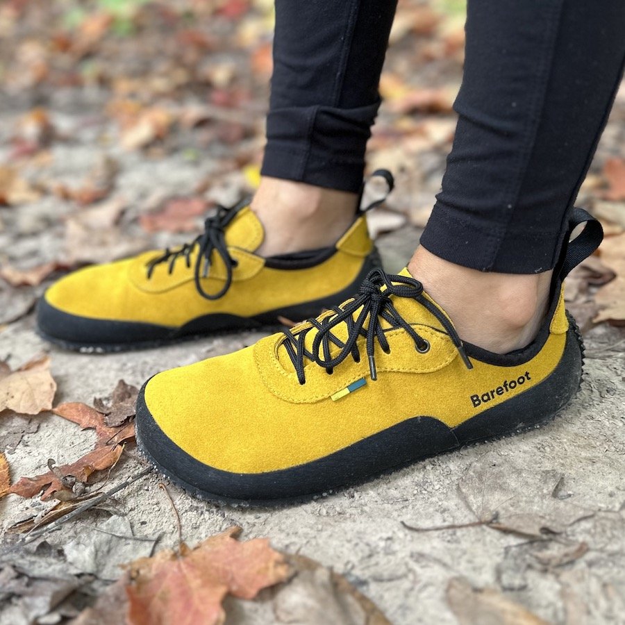 a close up side view of a person's feet wearing be lenka trailwalkers in mustard, the best barefoot hiking trail shoes, standing on a dirt path with fallen leaves around
