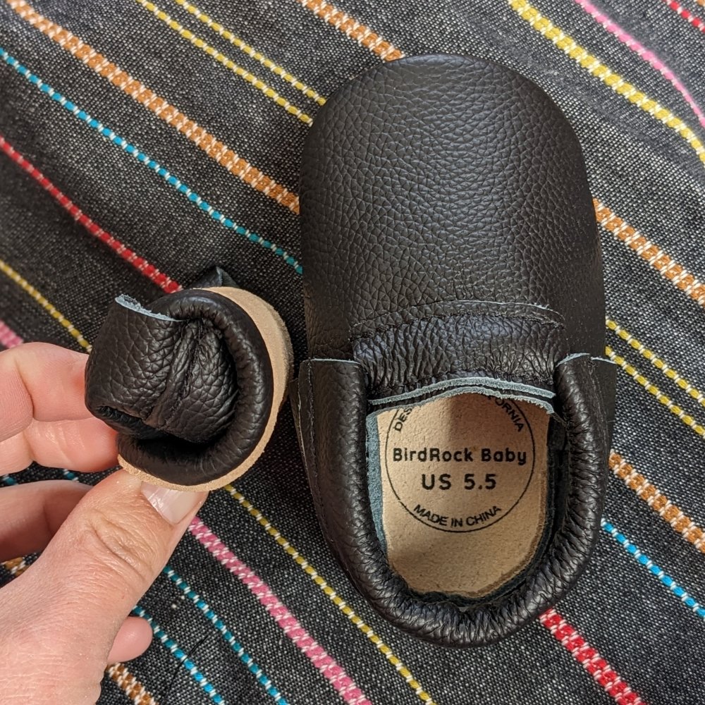 Bird Rock baby Moccasins with 1 shoe viewed from the top down in black leather and the other rolled into a tight ball