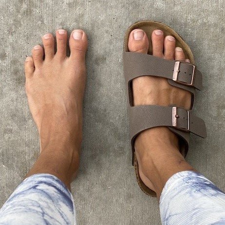 Top down view of a pair of feet wearing 1 Birkenstock Sandal and one foot bare to show the anatomical wide toe box fit