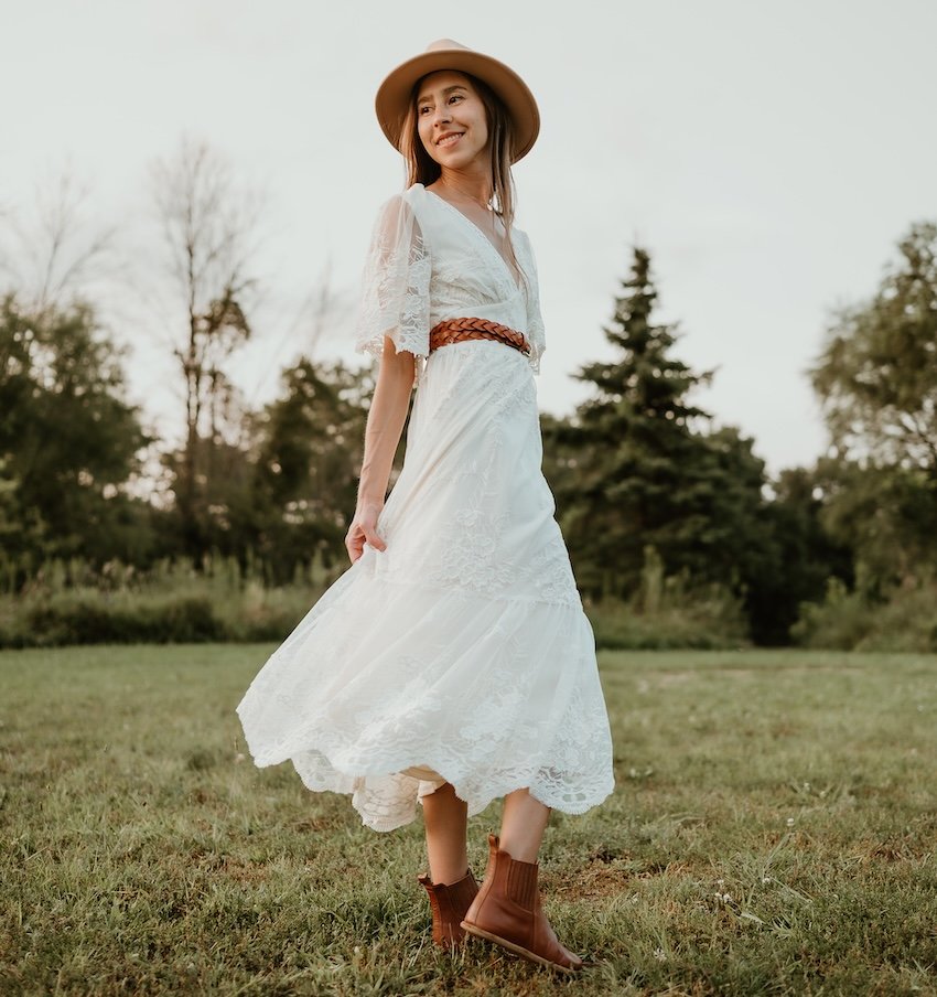 A full body view of a woman twirling in a lace dress in a field. She has accessorized her outfit with a tan hat, brown leather belt, and Anya's Shop Lila barefoot chelsea boots