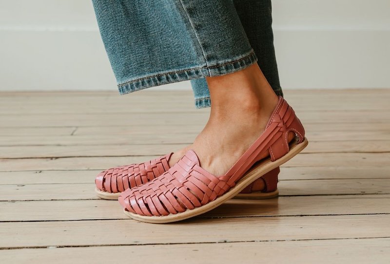 A close up left side view of a person's feet wearing the coral color Origo Huaraches, a leather woven styled flat. The person is wearing jeans and standing on a light wooden floor.