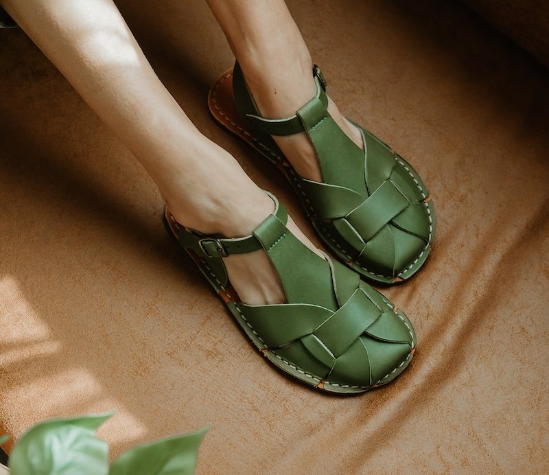 A close up of a woman's feet resting on a couch wearing foot shaped wide toe box Crupon Barefoot sandals Abbie, which is a leather fisherman style sandal, in olive green