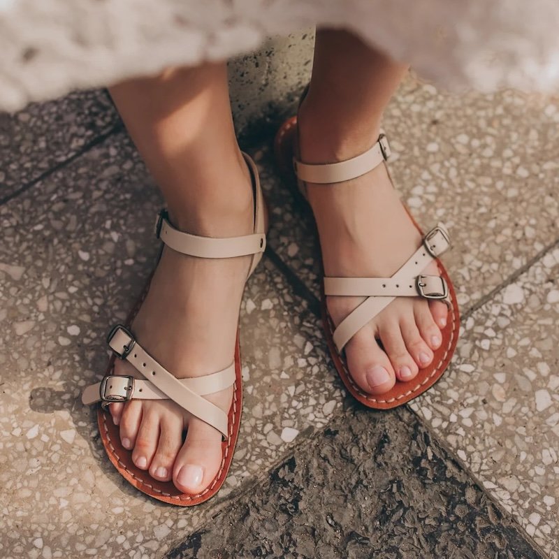 A top down view of a woman't feet with the hem of her white lace dress visible. She is wearing beige leather Crupon barefoot sandals with a cross strap. Model is called Norma.