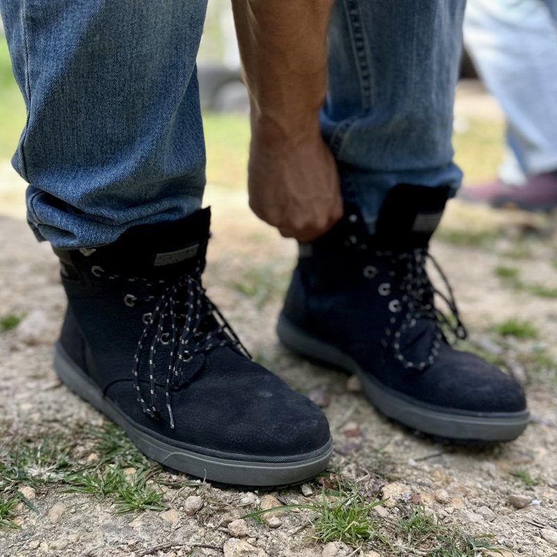 Close up view of a person putting DeWalt Plasma work boots on by grasping the boot shaft. The steel toe boot has a flat outsole and a wider than average toe box