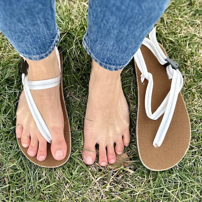 A top down view of a pair of feet standing on grass with one foot wearing a Earth Runners barefoot sandal Chronos with white Cloud laces, the other foot bare, and then the other Earth runners barefoot sandal sitting on the grass next to the bare foot