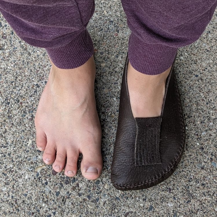 Top down close up of a woman with extra wide feet. One foot is bare, and the other is wearing a handmade leather moccasins from the Earthing Moccasins DIY tutorial and leather kit. It matches her foot shape perfectly